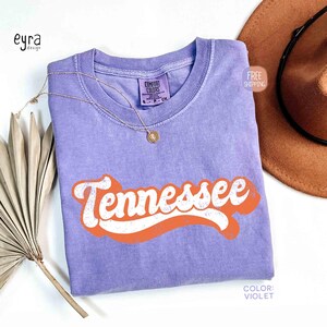 May include: A lilac Comfort Colors t-shirt featuring the word "Tennessee" in white with an orange outline. A gold necklace and a brown hat are also in the image. The shirt has a retro design.