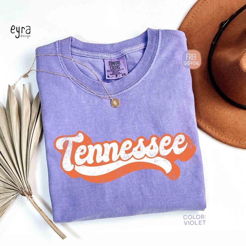 May include: A lilac Comfort Colors t-shirt featuring the word "Tennessee" in white with an orange outline. A gold necklace and a brown hat are also in the image. The shirt has a retro design.