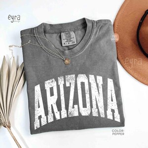 May include: A pepper-colored short-sleeve t-shirt featuring the word ARIZONA printed in a large, distressed white font across the chest. The shirt has a crew neckline and a Comfort Colors brand label on the inside collar. A thin gold necklace rests on the fabric.