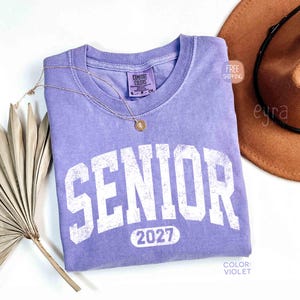 May include: A violet Comfort Colors t-shirt with the word "SENIOR" in large, distressed white letters and the year "2027" below. A gold necklace and a brown hat are next to the shirt. The shirt is folded on a white surface.
