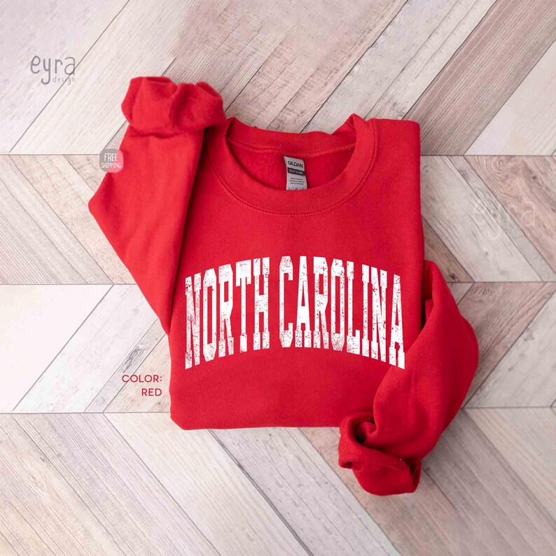 Może przedstawiać: Czerwona bluza z okrągłym dekoltem i napisem NORTH CAROLINA wykonanym przecieraną białą czcionką, umieszczonym łukiem na klatce piersiowej. Odzież posiada długie rękawy z prążkowanymi mankietami oraz prążkowany dekolt w klasycznym sportowym stylu.