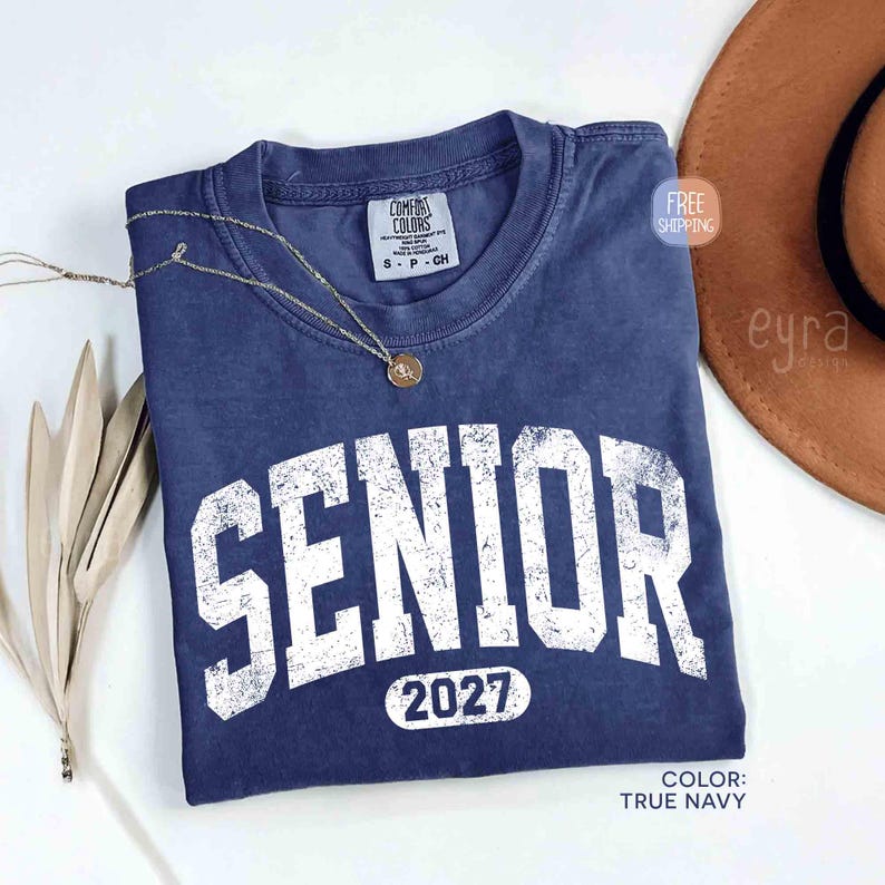 May include: A true navy blue t-shirt with the word "SENIOR" in distressed white lettering and the year "2027" below. A gold necklace rests on the shirt. The shirt is from Comfort Colors.