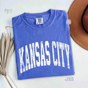 May include: A folded, periwinkle blue t-shirt with the text "KANSAS CITY" in distressed white lettering. The shirt has a crew neck and a tag at the neckline. The color is described as "FLO BLUE".