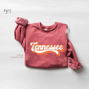 K&ouml;nnte beinhalten: Ein scharlachrotes Langarm-Sweatshirt mit Rundhalsausschnitt, das das Wort Tennessee in einer wei&szlig;en, retro-artigen Schreibschrift &uuml;ber einer einfarbigen, orangefarbenen Wellenform zeigt. Das Kleidungsst&uuml;ck hat eine strukturierte Stoffoptik.