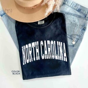 K&ouml;nnte beinhalten: Ein schwarzes T-Shirt mit einem verwaschenen, wei&szlig;en, bogenf&ouml;rmigen Schriftzug, der NORTH CAROLINA lautet. Das T-Shirt liegt unter einem beigen Hut mit breiter Krempe und einer hellblauen Jeans, mit einer kleinen goldenen Halskette auf dem Stoff.