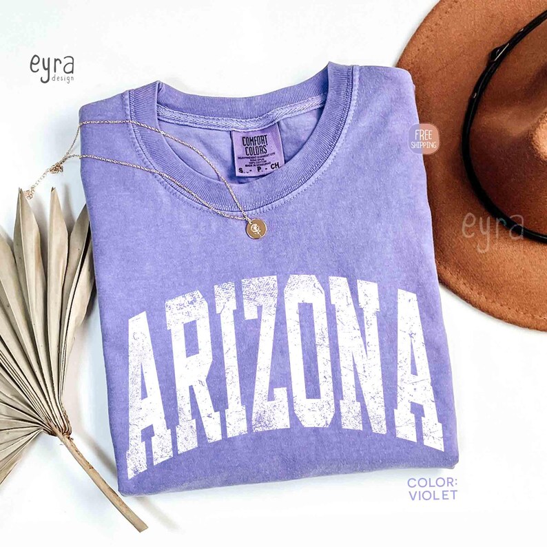 May include: A violet short-sleeved t-shirt with a crew neckline featuring the word ARIZONA printed in large, distressed white capital letters across the chest. The shirt has a Comfort Colors brand label at the inner neck and a thin gold necklace.