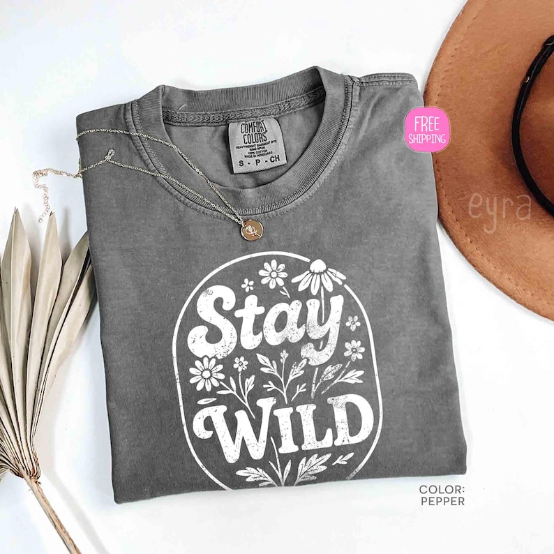 May include: A gray Comfort Colors t-shirt with the words "Stay Wild" in a white floral design. A gold necklace rests on the shirt. The shirt is labeled "Pepper" in color.
