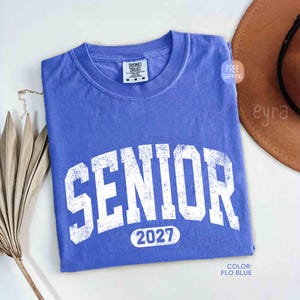 May include: A blue t-shirt with the word "SENIOR" in large, distressed white letters. Below the word "SENIOR" is the number "2027". The shirt is folded and has a crew neck. The color is "FLO BLUE".