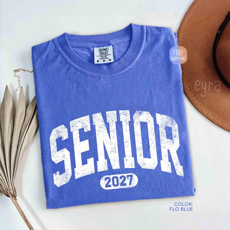 May include: A blue t-shirt with the word "SENIOR" in large, distressed white letters. Below the word "SENIOR" is the number "2027". The shirt is folded and has a crew neck. The color is "FLO BLUE".