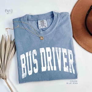 Könnte beinhalten: Ein kurzärmeliges T-Shirt in Jeansblau mit Rundhalsausschnitt. Das Shirt zeigt den Schriftzug BUS DRIVER in einer abgenutzten weißen Schrift auf der Brust. Eine goldfarbene Halskette mit einem kleinen runden Anhänger liegt am Kragenbereich.