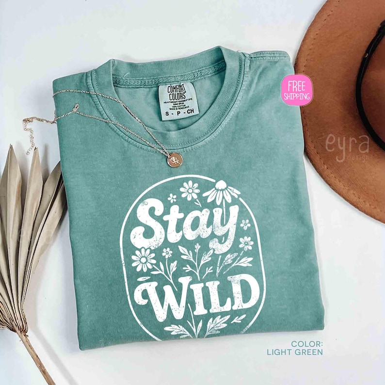 May include: Light green t-shirt with the text "Stay Wild" in a white floral design. The shirt is a Comfort Colors brand, size S-P-CH. A silver necklace with a pendant is on the shirt. The color is light green.