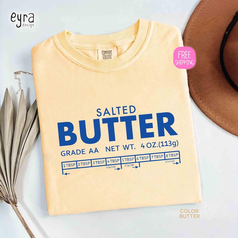 May include: A light yellow t-shirt with the word "BUTTER" in large blue letters. The text "SALTED" is above the word "BUTTER". Below the word "BUTTER" is the text "GRADE AA NET WT. 4 OZ.(113g)". There is a measurement chart below the text.