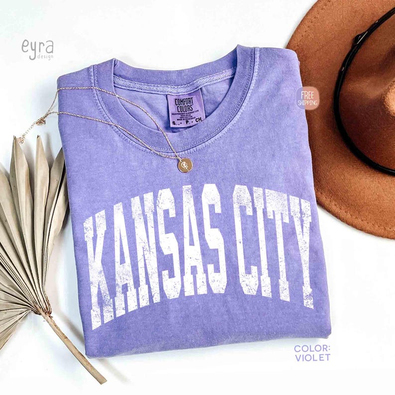 May include: A violet-colored t-shirt with the text "KANSAS CITY" in white, arched lettering. A gold necklace with a pendant rests on the shirt. A brown hat is visible in the background. The shirt is labeled "Comfort Colors".