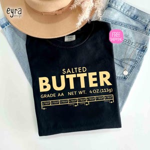 May include: Black t-shirt with the words "SALTED BUTTER" in gold lettering. The shirt also features a butter measurement guide and the text "GRADE AA NET WT. 4 OZ. (113g)". A tan hat and blue jeans are in the background.