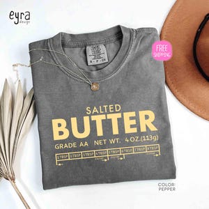 May include: A gray t-shirt with the words "SALTED BUTTER" in yellow block letters. Below the text is "GRADE AA NET WT. 4 OZ. (113g)" and a measurement guide. A silver necklace with a pendant rests on the shirt.