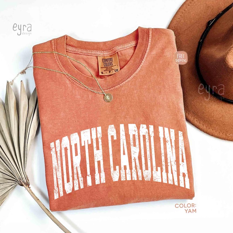 K&ouml;nnte beinhalten: Ein terrakottafarbenes Kurzarm-T-Shirt mit Rundhalsausschnitt und einem verwaschenen wei&szlig;en Grafikdruck, der in geschwungener Schrift NORTH CAROLINA zeigt. Das T-Shirt hat ein Comfort Colors-Etikett am Innenhals und eine goldfarbene Halskette.
