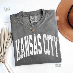 May include: A gray Comfort Colors t-shirt with the text "KANSAS CITY" in distressed white lettering. A gold necklace and a brown hat are next to the shirt. The shirt is labeled "Pepper" in color.