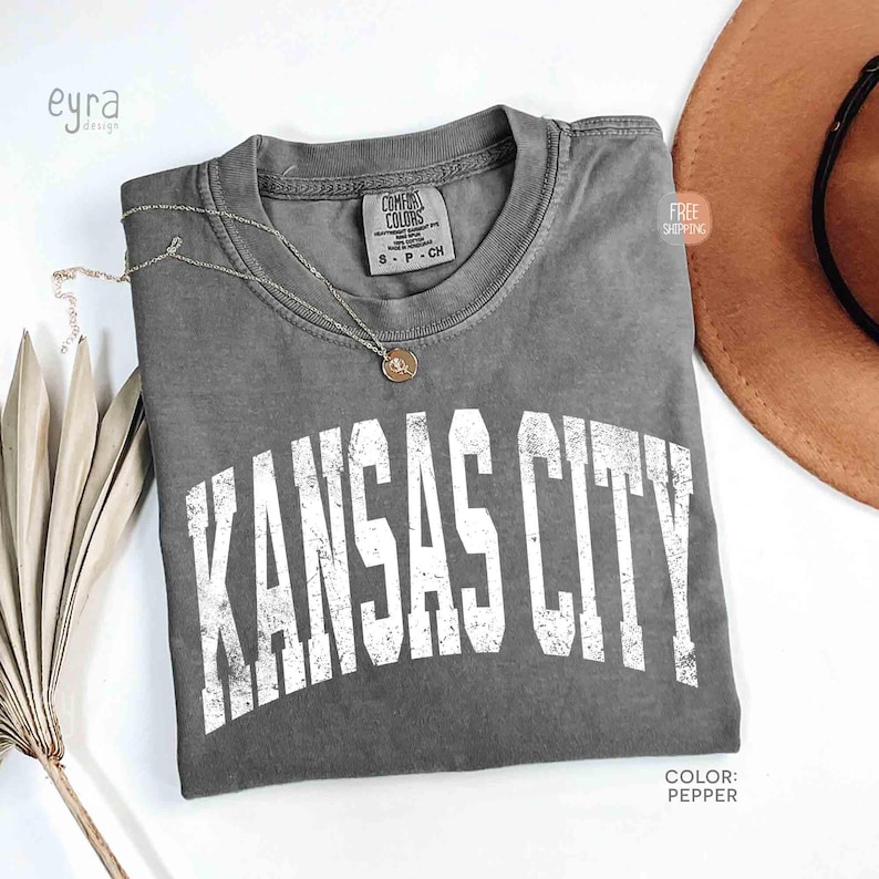 May include: A gray Comfort Colors t-shirt with the text "KANSAS CITY" in distressed white lettering. A gold necklace and a brown hat are next to the shirt. The shirt is labeled "Pepper" in color.