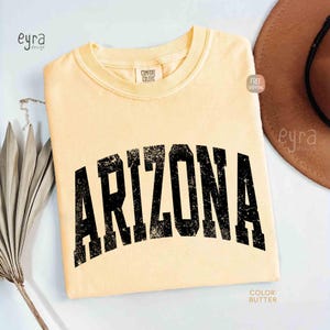 May include: A butter-colored short-sleeve t-shirt featuring the word ARIZONA printed in a large, distressed black collegiate-style font across the chest. The shirt has a crew neckline and a Comfort Colors brand label visible at the inner collar.