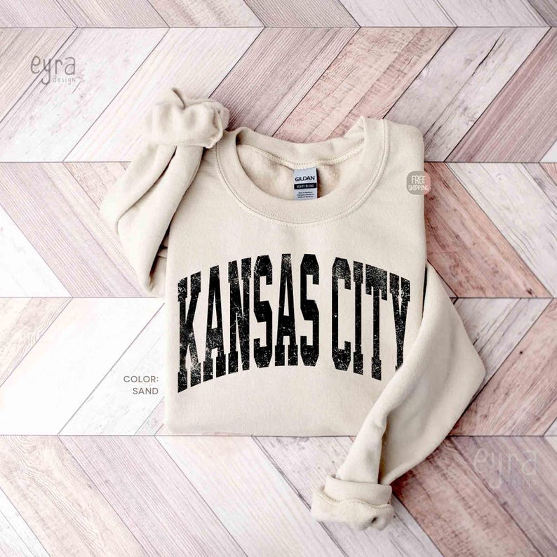 May include: A sand-coloured sweatshirt featuring the text "KANSAS CITY" in a distressed black font. The sweatshirt is folded, with rolled-up sleeves. The colour is labelled "SAND".