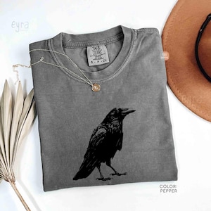 May include: A pepper-colored crew neck t-shirt featuring a black graphic illustration of a raven centered on the chest. The shirt has a visible interior neck label that reads Comfort Colors, 100% cotton, made in Honduras, and size S P CH.