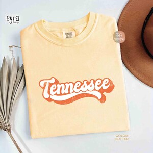 May include: Pale yellow t-shirt featuring the word "Tennessee" in a retro orange and white design. The shirt is a Comfort Colors brand. A brown hat is partially visible in the background.