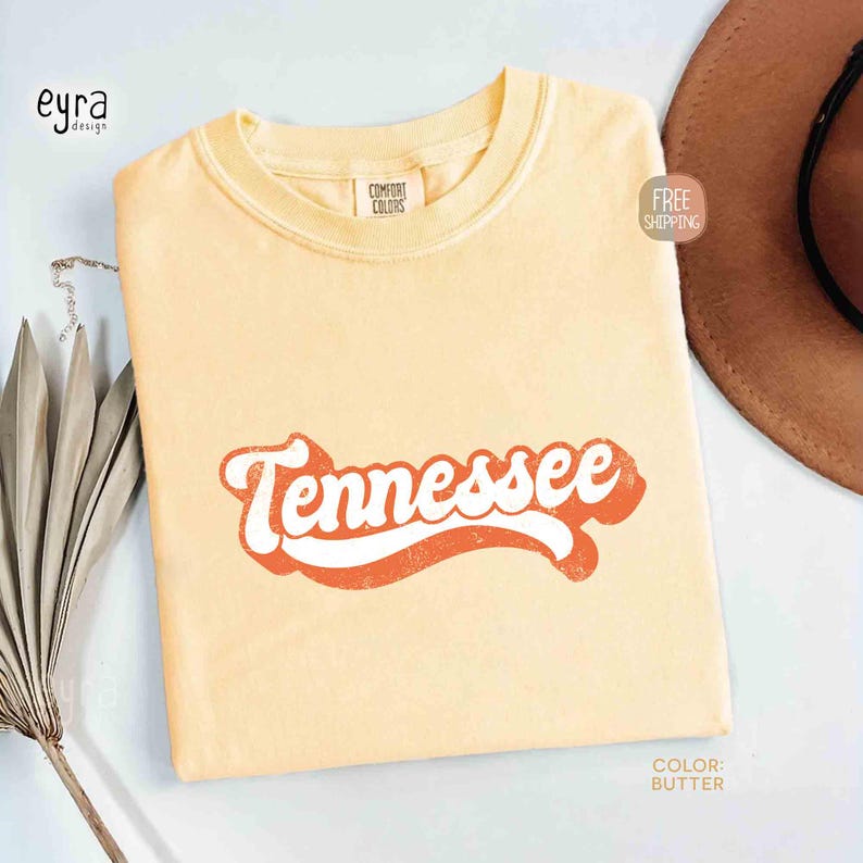May include: Pale yellow t-shirt featuring the word "Tennessee" in a retro orange and white design. The shirt is a Comfort Colors brand. A brown hat is partially visible in the background.