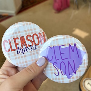 May include: Two circular pin-back buttons featuring a light orange and white plaid pattern. One button displays the text CLEMSON tigers in orange and purple. The other button features the text CLEM SON in purple over a purple state silhouette.