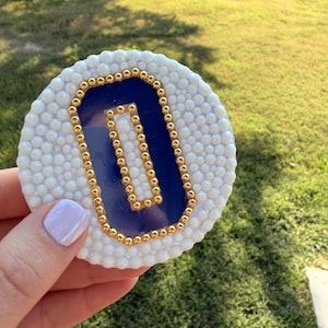May include: A circular coaster featuring a white textured border made of small raised beads surrounding a smooth, deep blue centre. A rectangular frame composed of small, metallic gold beads is centred on the blue surface, creating a decorative accent.