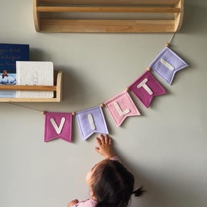 Può includere: Una ghirlanda di feltro con la scritta "V I L L T" in lettere bianche su bandierine rosa e viola. La ghirlanda è infilata con perline di legno e appesa a una parete verde chiaro. Un bambino allunga la mano verso la ghirlanda.