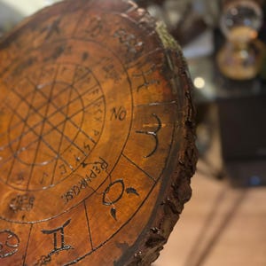 May include: A round, wooden table top with a dark brown finish. The surface is etched with astrological symbols and the word "REPHRASE". The edge of the table has a natural, rough bark texture.