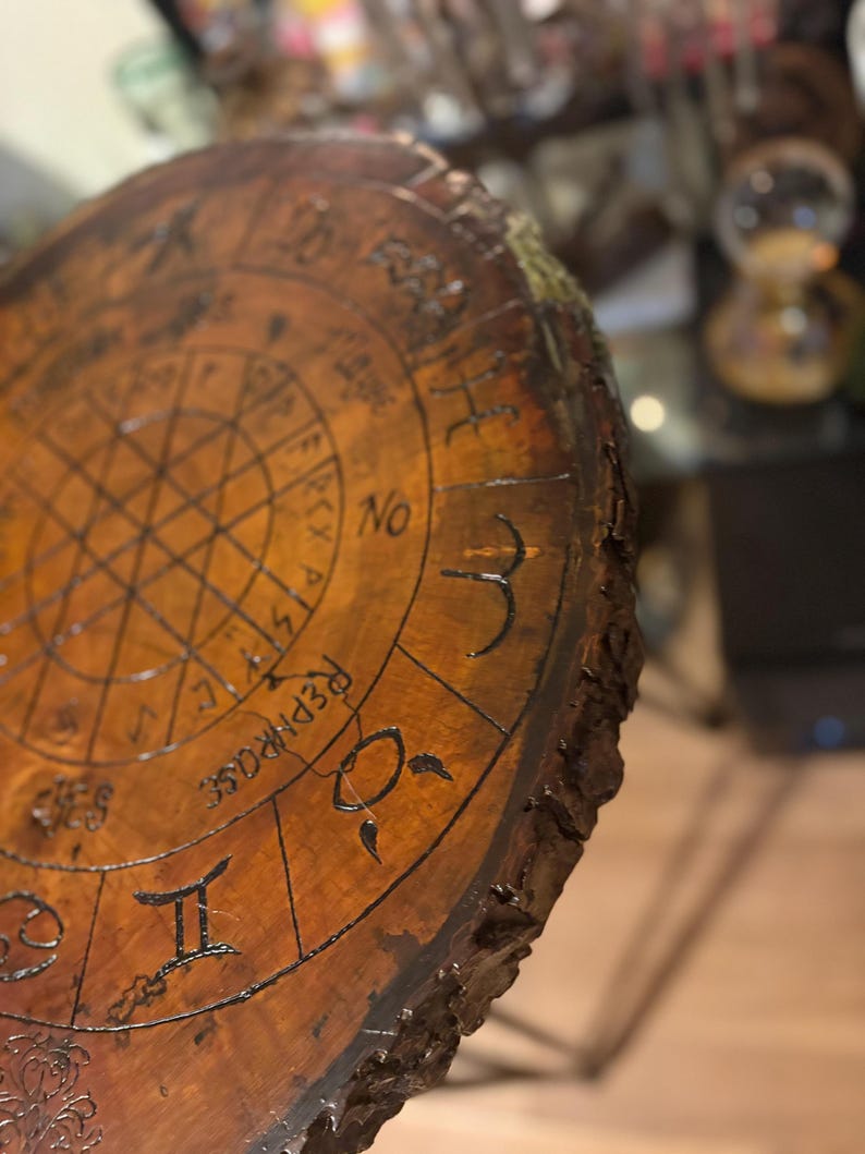 May include: A round, wooden table top with a dark brown finish. The surface is etched with astrological symbols and the word "REPHRASE". The edge of the table has a natural, rough bark texture.