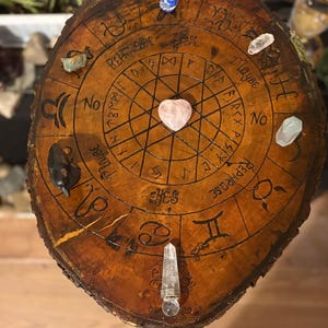 May include: A round, wooden divination board with a dark brown finish. The board features etched zodiac symbols, runes, and the words "Yes," "No," and "Maybe." Various crystals, including a heart-shaped rose quartz, are placed around the board.