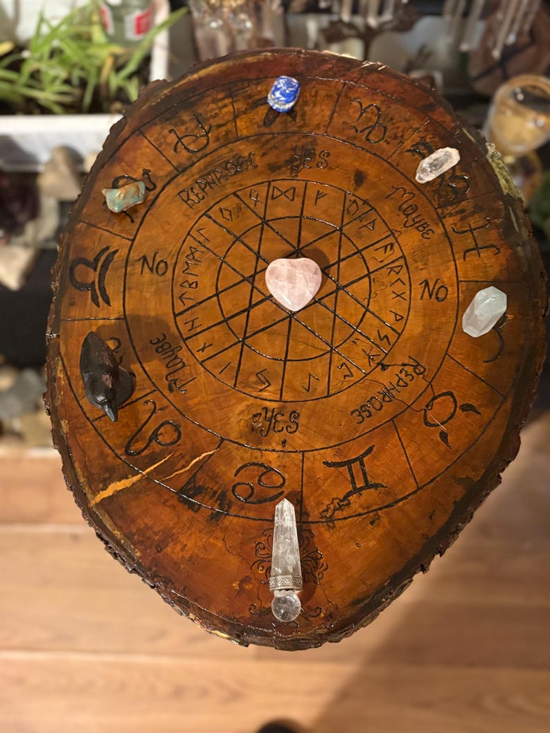 May include: A round, wooden divination board with a dark brown finish. The board features etched zodiac symbols, runes, and the words "Yes," "No," and "Maybe." Various crystals, including a heart-shaped rose quartz, are placed around the board.