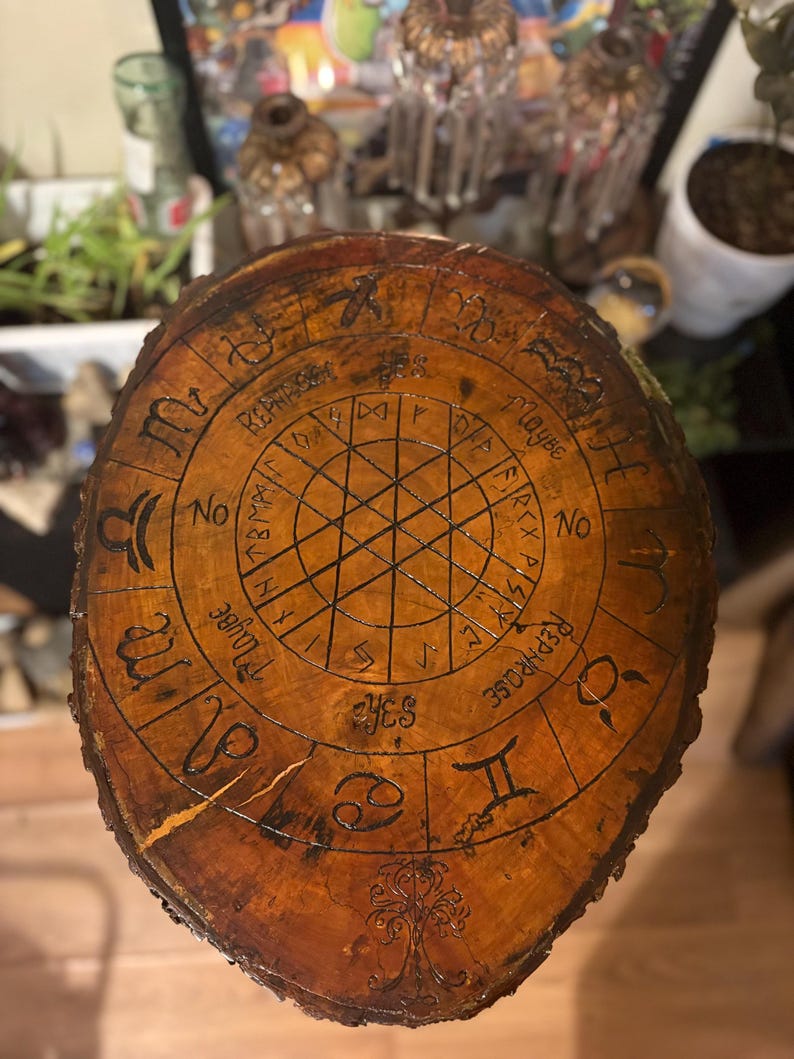 May include: A round, wooden divination board with a dark brown finish. The board features etched zodiac symbols, runes, and the words "Yes", "No", and "Maybe". A tree design is at the bottom.