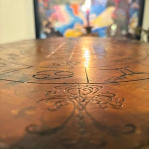 May include: A close-up of a round, copper-colored table with etched zodiac symbols and a central tree-of-life design. A lit candle is in the background, casting a warm glow. The table's surface is detailed with lines and patterns.