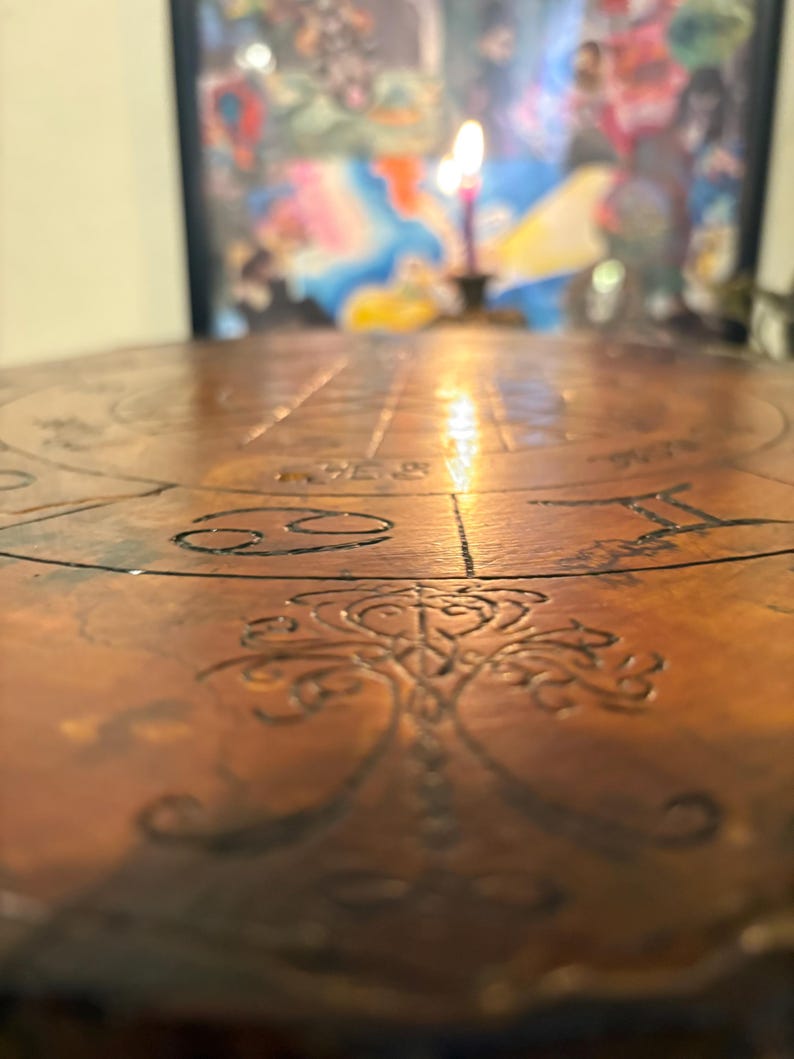 May include: A close-up of a round, copper-colored table with etched zodiac symbols and a central tree-of-life design. A lit candle is in the background, casting a warm glow. The table's surface is detailed with lines and patterns.