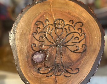 Pode incluir: Uma fatia circular de madeira com um desenho de linhas escuras queimadas de uma árvore com um símbolo circular no topo. Um pequeno cristal de ametista áspero e de cor violeta clara está incrustado nos veios da madeira à esquerda.