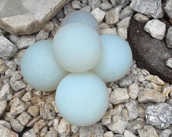 May include: A cluster of five pale blue glass spheres, each about 3 inches in diameter, resting on a bed of mixed gray and tan gravel. A snail shell sits on a light-colored stone in the background.