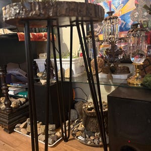 May include: A small, round side table with a natural wood slab top and black hairpin legs. The wood top has a rough, textured edge. The legs are thin and angled, providing a modern, minimalist aesthetic. The table is suitable for indoor use.