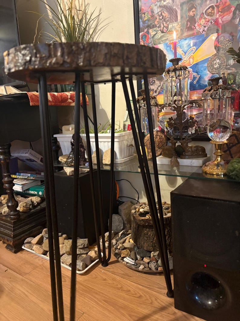 May include: A small, round side table with a natural wood slab top and black hairpin legs. The wood top has a rough, textured edge. The legs are thin and angled, providing a modern, minimalist aesthetic. The table is suitable for indoor use.