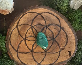 May include: A green malachite gemstone pendant rests on a wooden slice with a carved Seed of Life design. The wood has a natural, warm brown tone with a dark, etched pattern. The background includes moss and crystals.