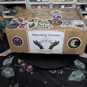 May include: A brown cardboard box covered in various stickers featuring celestial, skeletal, and book-themed illustrations. A central white label displays the text Mystery Coven, an illustration of two hands holding a crystal, and Stationery bundles and witchy delights.