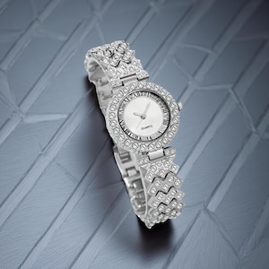 May include: Silver-toned quartz watch with a round face and a band adorned with sparkling crystals. The watch face features Roman numerals and the word "QUARTZ". The watch is displayed on a textured, gray surface.