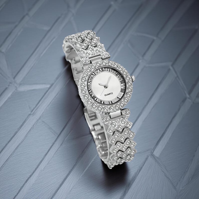 May include: Silver-toned quartz watch with a round face and a band adorned with sparkling crystals. The watch face features Roman numerals and the word "QUARTZ". The watch is displayed on a textured, gray surface.