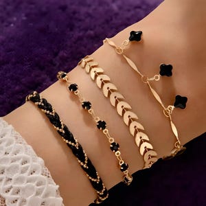 May include: Four gold-coloured bracelets are shown on a wrist. One is a braided black cord, another features black gemstones, a third has a leaf design, and the last has black clover charms. The bracelets are delicate and elegant.