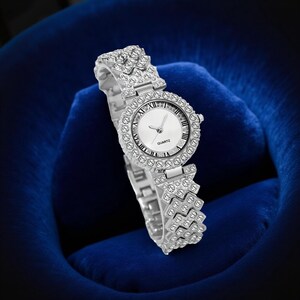 May include: Silver-toned quartz watch with a round face and a bezel and band encrusted with small, sparkling crystals. The watch face is white with black Roman numerals and the word "QUARTZ" printed at the bottom. The watch is displayed on a blue velvet surface.