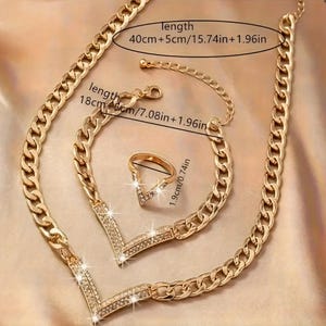 以下が含まれることがあります： ゴールドカラーのジュエリーセット。ネックレス、ブレスレット、リングが含まれています。ネックレスとブレスレットは、小さなクリアストーンで飾られたV字型のペンダントが付いたチェーンデザインです。リングも同様の石を使用したV字型デザインです。