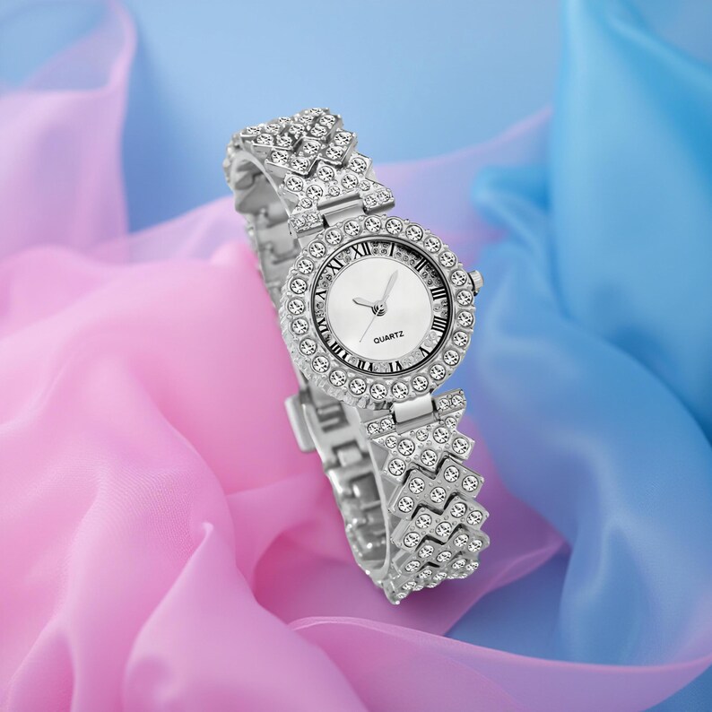 May include: A silver-toned quartz watch with a round face and a band adorned with sparkling crystals. The watch face is white with black Roman numerals and hands. The word "QUARTZ" is printed on the watch face.