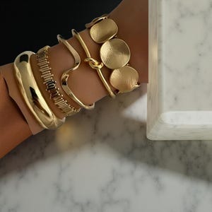 May include: A selection of gold-coloured bracelets. The bracelets include a thick, rounded bangle, a textured band, a wavy design, a knot-style bracelet, and a bracelet with three circular, textured charms. The bracelets are displayed on a wrist.